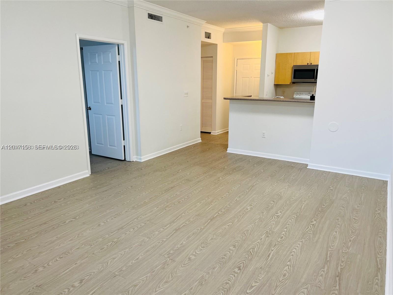An empty living room with light wood-look flooring leads to an open kitchen with cabinets — 1900 Van Buren St # 111B, Hollywood FL 33020, HOLLYWOOD, FL