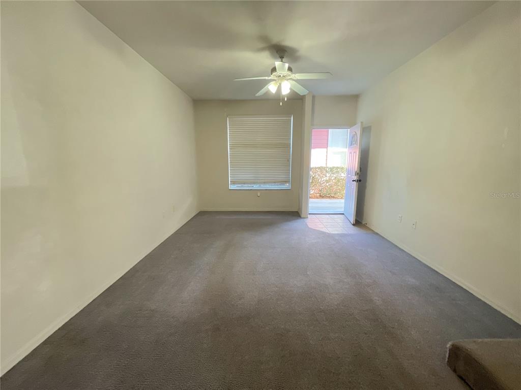 An empty living room features beige carpet, a ceiling fan, a window, and an open door — 3930 SW 20TH AVENUE UNIT 1902, GAINESVILLE, FL