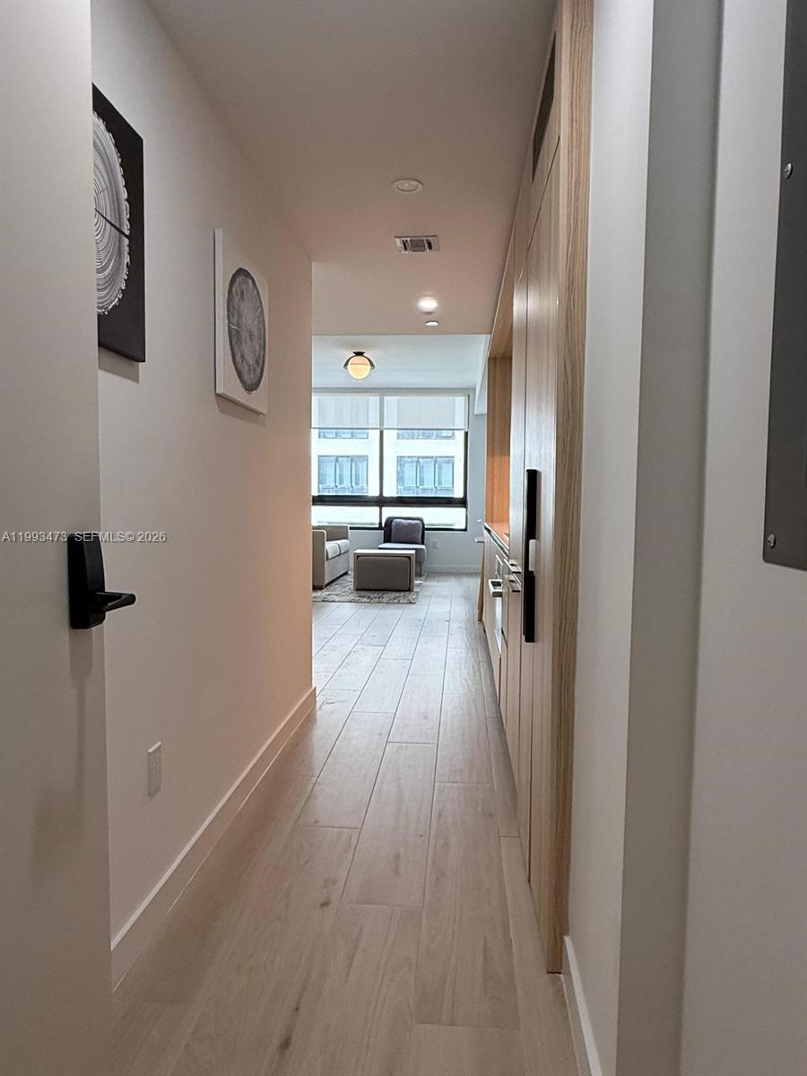 A narrow hallway with light wood flooring leads toward a bright living area with windows — 225 N Miami Ave # 910, Miami FL 33128, MIAMI, FL