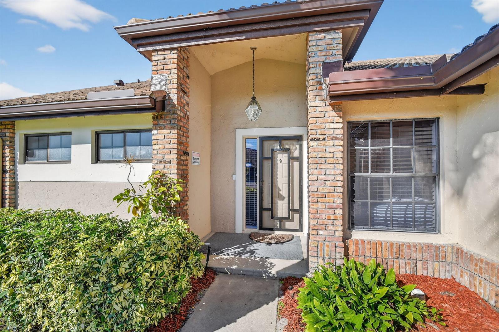 The exterior of a house features a brick-trimmed entryway, a glass storm door, and landscaping — 7147 Le Chalet Boulevard, Boynton Beach FL 33472, BOYNTON BEACH, FL