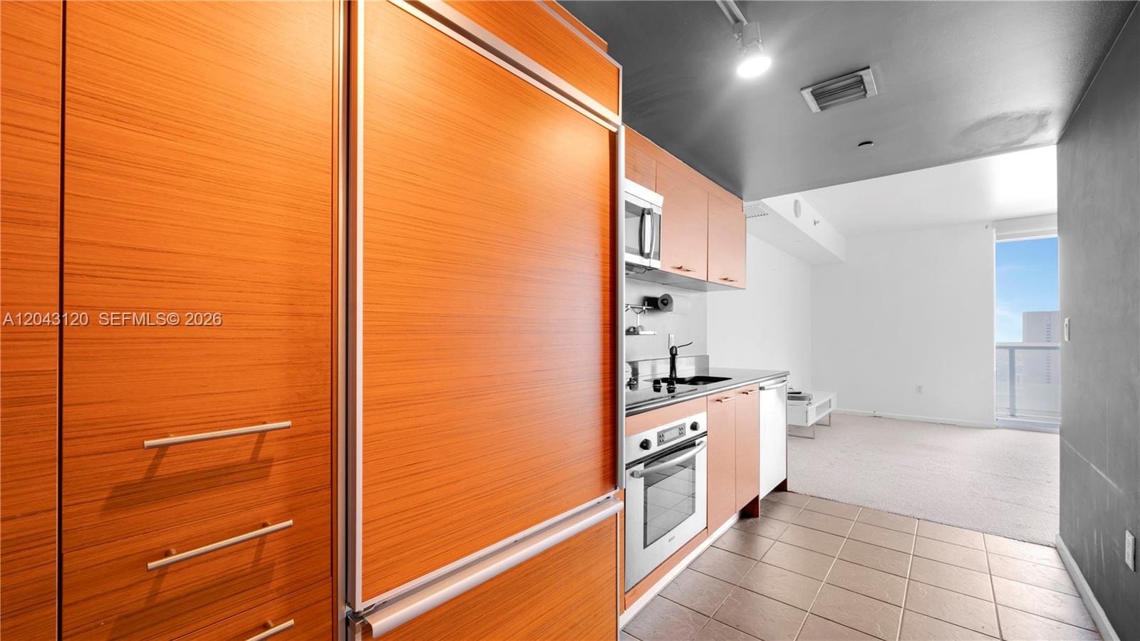 A kitchen with wood-grain cabinetry and stainless steel appliances opens into a carpeted living area — 50 Biscayne Blvd # 4203, Miami FL 33132, MIAMI, FL