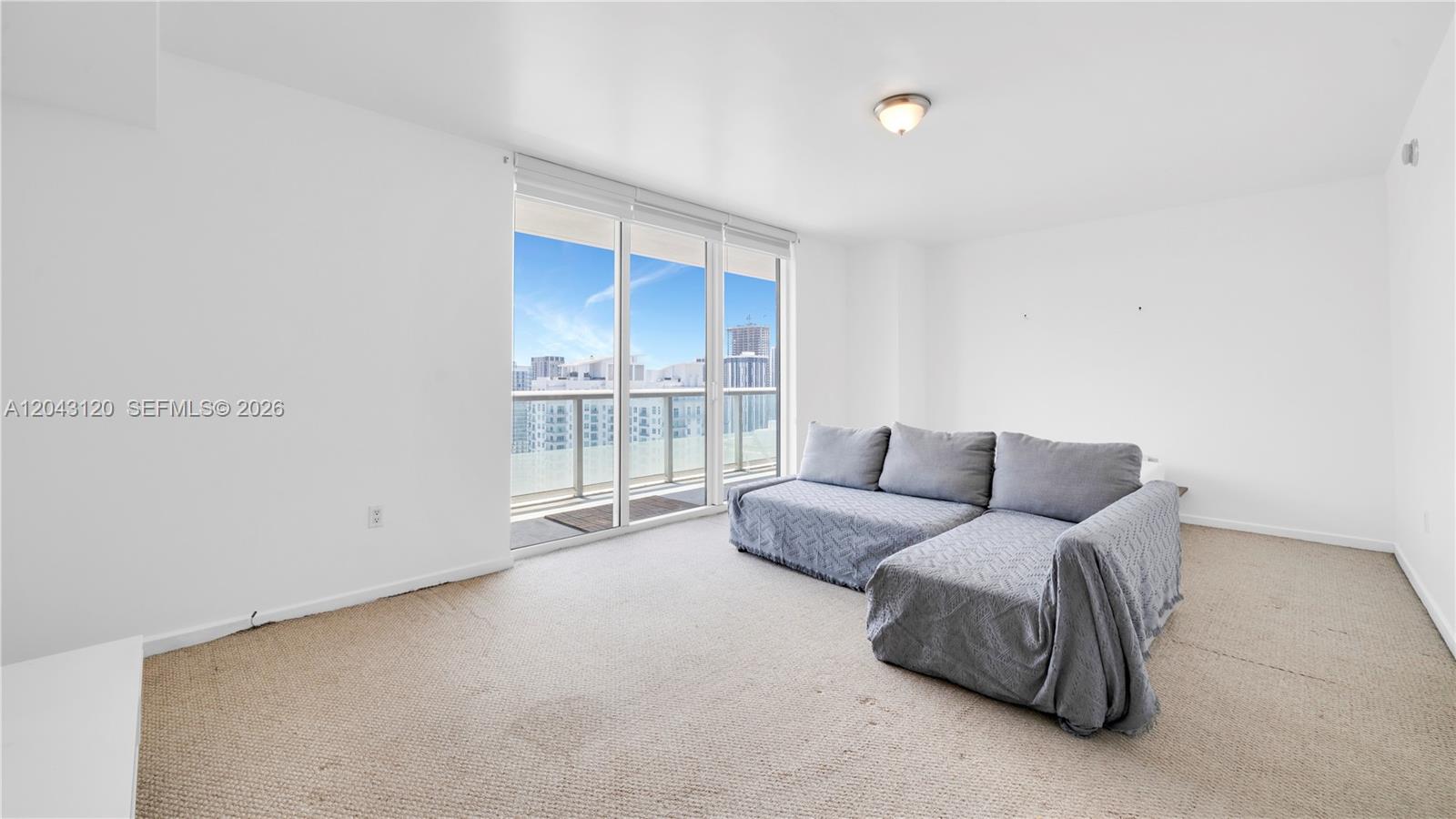 A living room features beige carpeting, white walls, a gray sectional sofa, and balcony access — 50 Biscayne Blvd # 4203, Miami FL 33132, MIAMI, FL