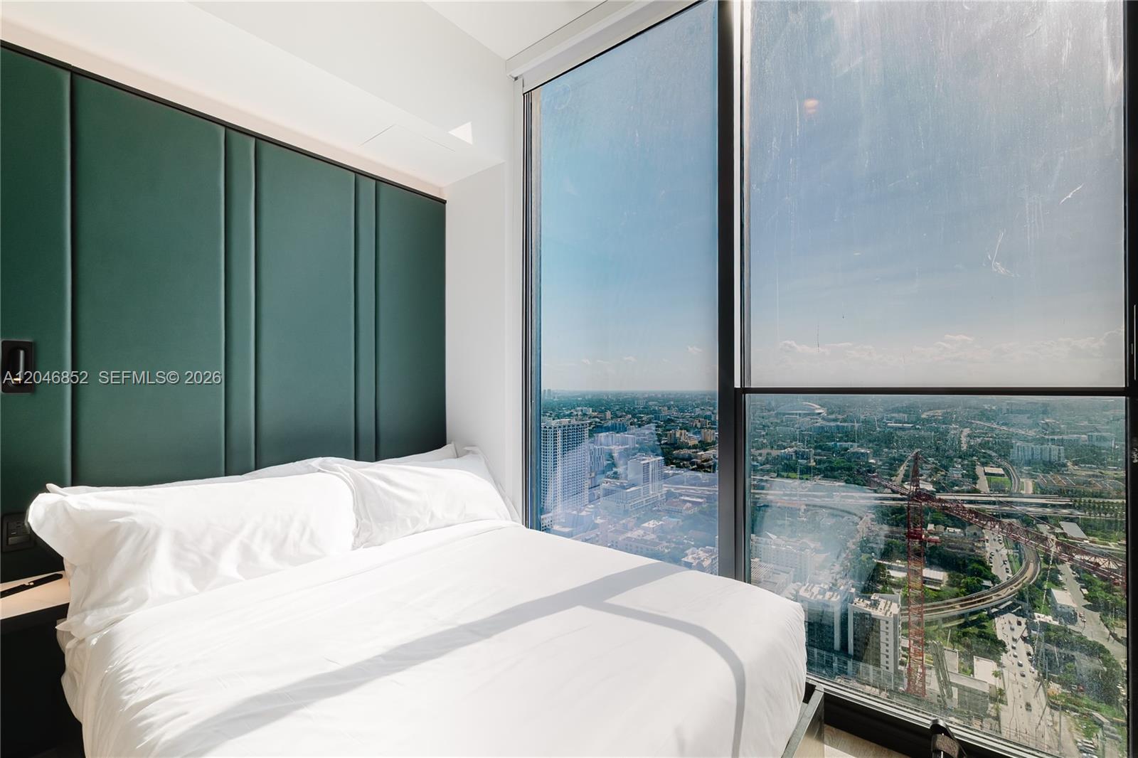 A bedroom features a green paneled headboard and floor-to-ceiling windows overlooking a city skyline — 20 NE 11th st # 4907A, Miami FL 33132, MIAMI, FL