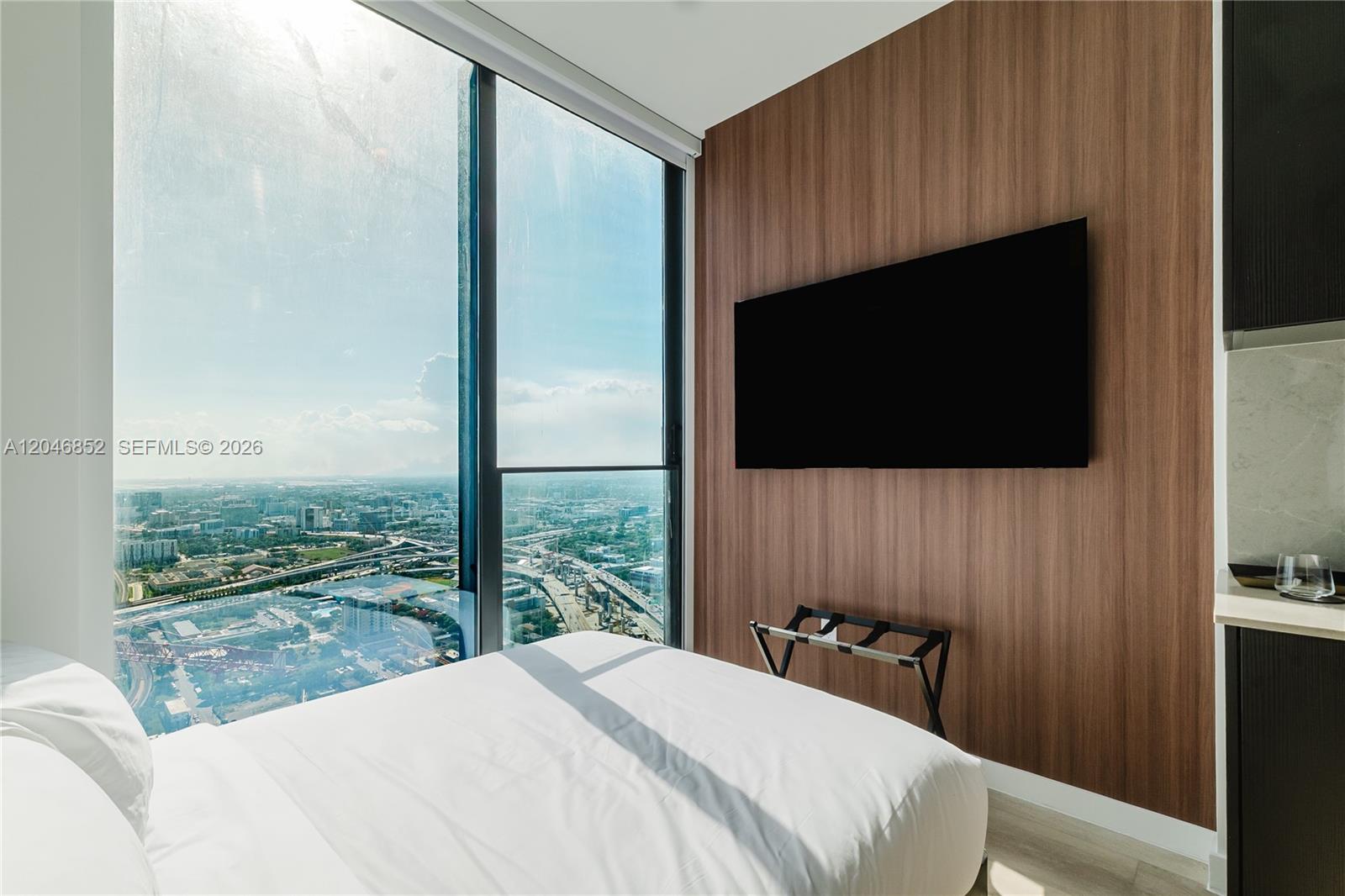 A bedroom features a wall-mounted television, a luggage rack, and floor-to-ceiling windows overlooking a city — 20 NE 11th st # 4907A, Miami FL 33132, MIAMI, FL
