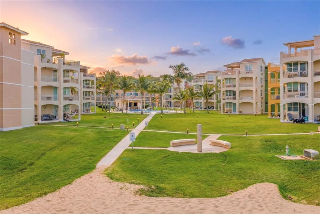 A grassy courtyard with palm trees and a walkway sits between multi-story apartment buildings — Haudimar Beach Apartments 4466 UNIT F208, ISABELA, PR