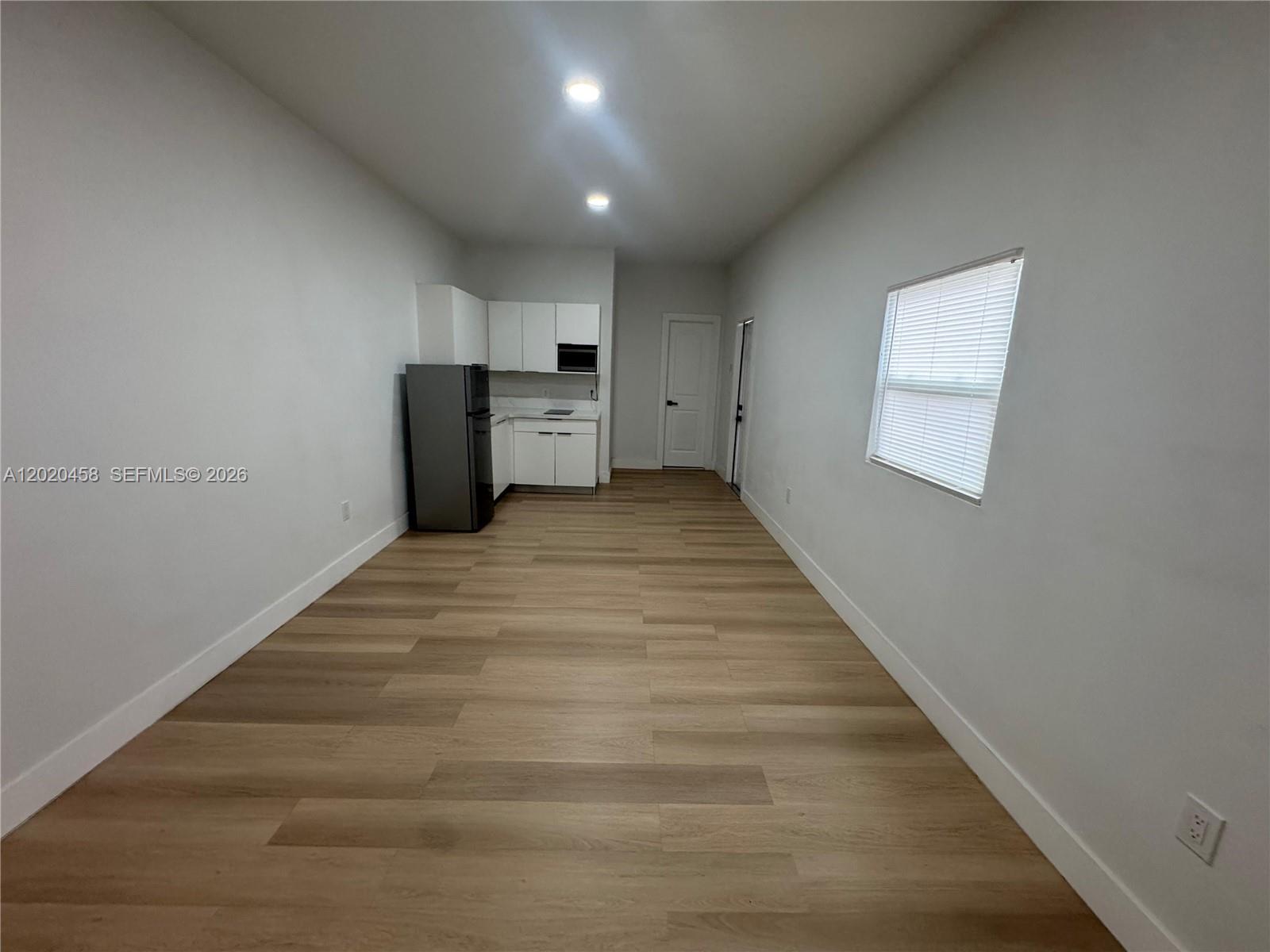 A long, narrow room features light wood-look flooring, white walls, and a small kitchen area — MIAMI, FL