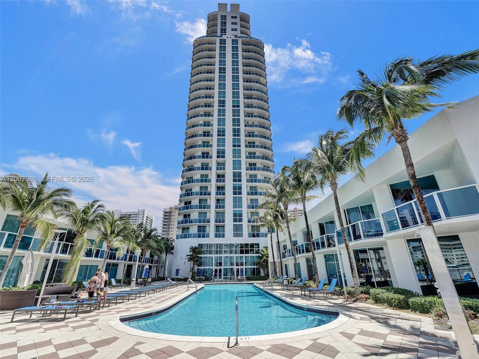 A tall residential tower rises above a swimming pool surrounded by palm trees and buildings — 1945 S Ocean Dr # 2101, Hallandale Beach FL 33009, HALLANDALE BEACH, FL