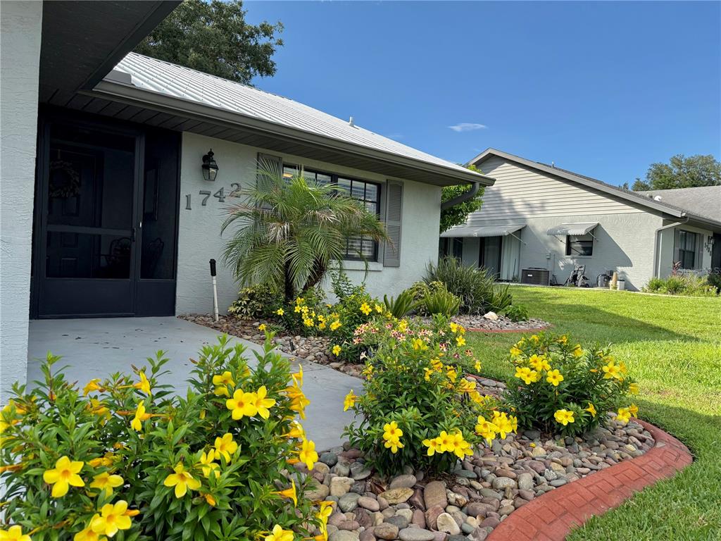 A single-story house exterior features yellow flowering shrubs, a rock garden, and a screened entryway — 1742 W ORANGEWOOD PLACE, AVON PARK, FL