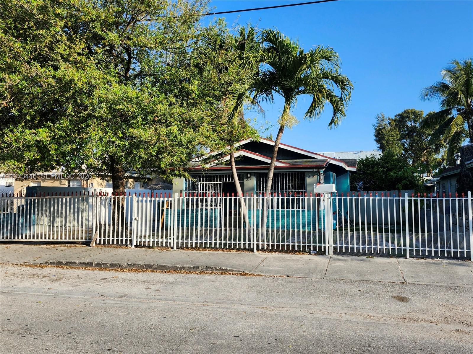 A single-story house with a blue exterior and a white metal fence sits behind palm trees — 227 NW 33rd St, Miami FL 33127, MIAMI, FL