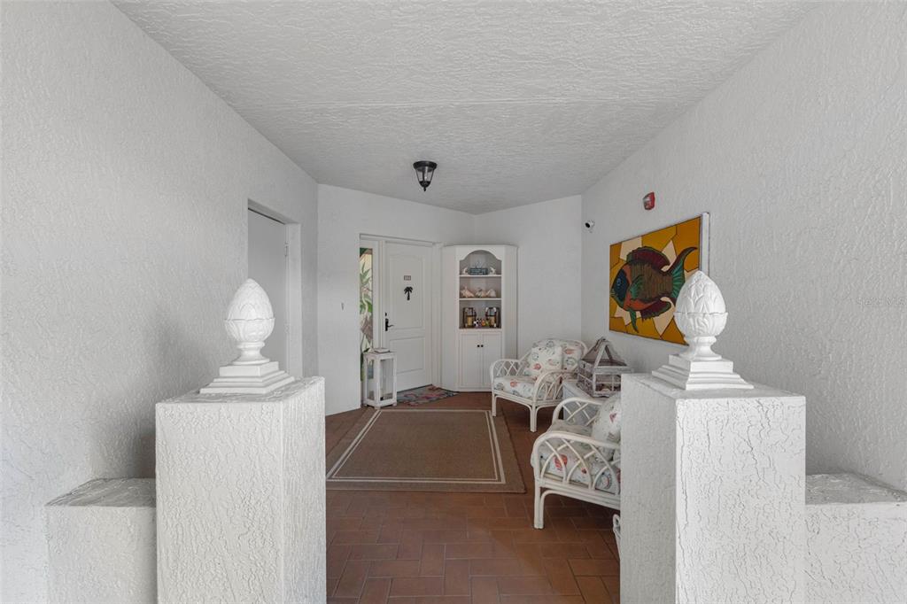 A covered entryway features white stucco walls, tiled flooring, two wicker chairs, and decorative finials — 200 121ST AVENUE UNIT 104, TREASURE ISLAND, FL