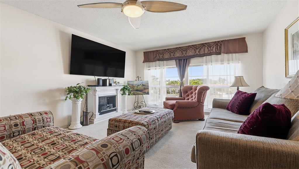 A living room features a fireplace, a large television, patterned furniture, and a ceiling fan — 2700 BAYSHORE BOULEVARD UNIT 1307, DUNEDIN, FL