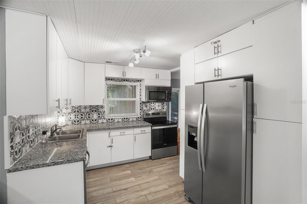 A kitchen features white cabinets, granite countertops, patterned tile backsplashes, and stainless steel appliances — 1663 SAN MATEO DRIVE, DUNEDIN, FL