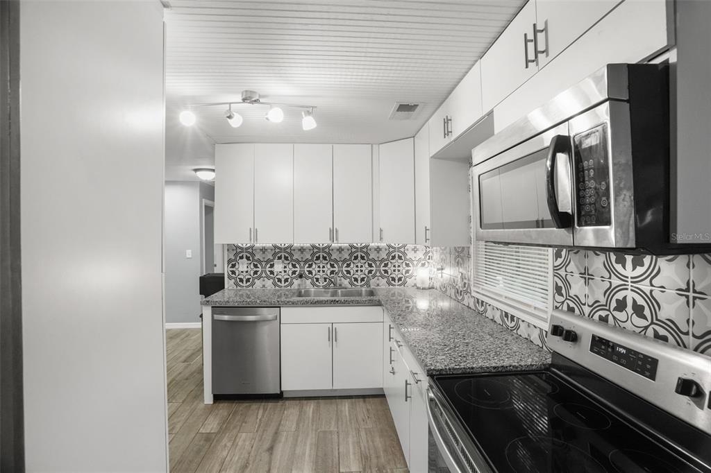 A kitchen features white cabinets, granite countertops, a patterned tile backsplash, and stainless steel appliances — 1663 SAN MATEO DRIVE, DUNEDIN, FL
