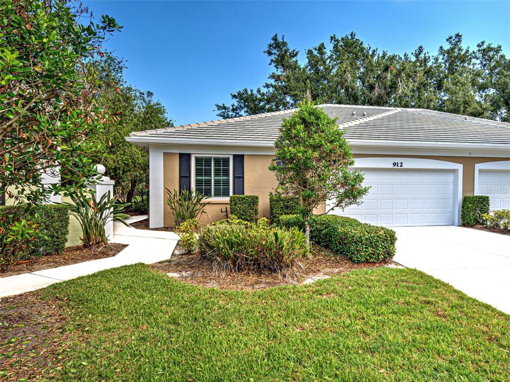 A single-story house exterior features a white garage door, tan walls, and manicured landscaping — 912 BARCLAY COURT, VENICE, FL