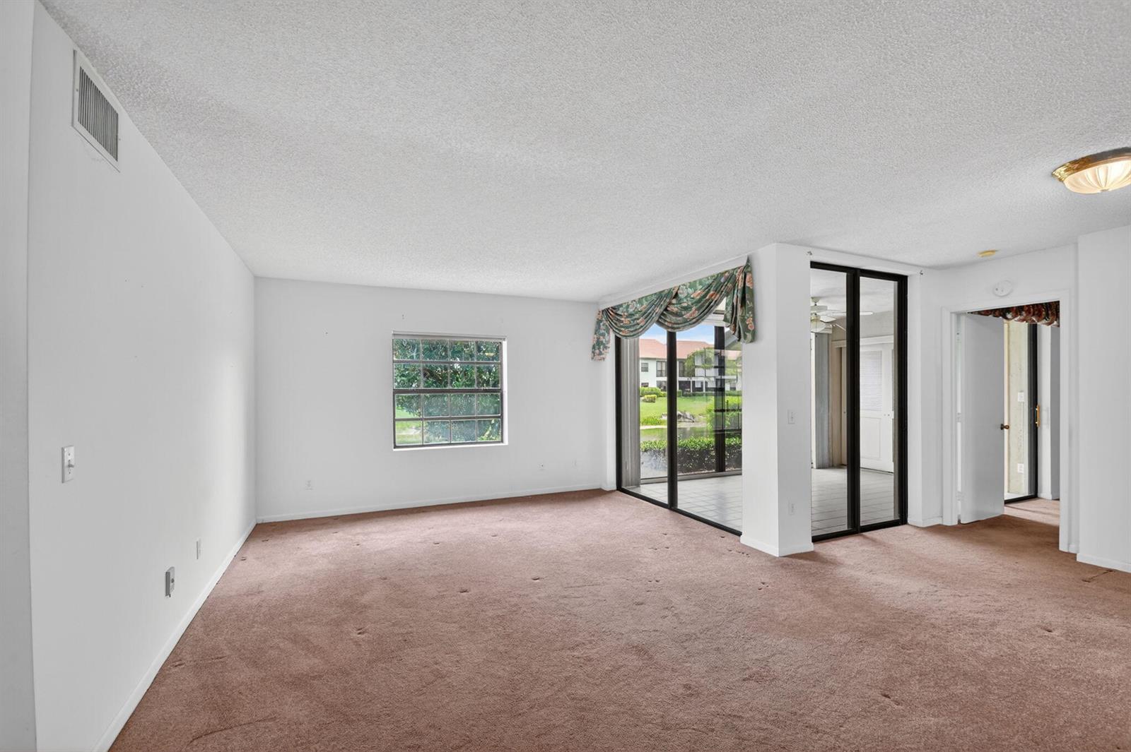 An empty living room features brown carpet, white walls, a window, and a glass-enclosed patio — 5395 Firenze Drive # B, Boynton Beach FL 33437, BOYNTON BEACH, FL