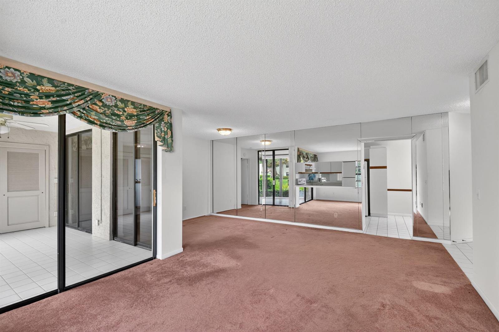 A living room with brown carpet, a mirrored wall, and a sliding glass door to a patio — 5395 Firenze Drive # B, Boynton Beach FL 33437, BOYNTON BEACH, FL