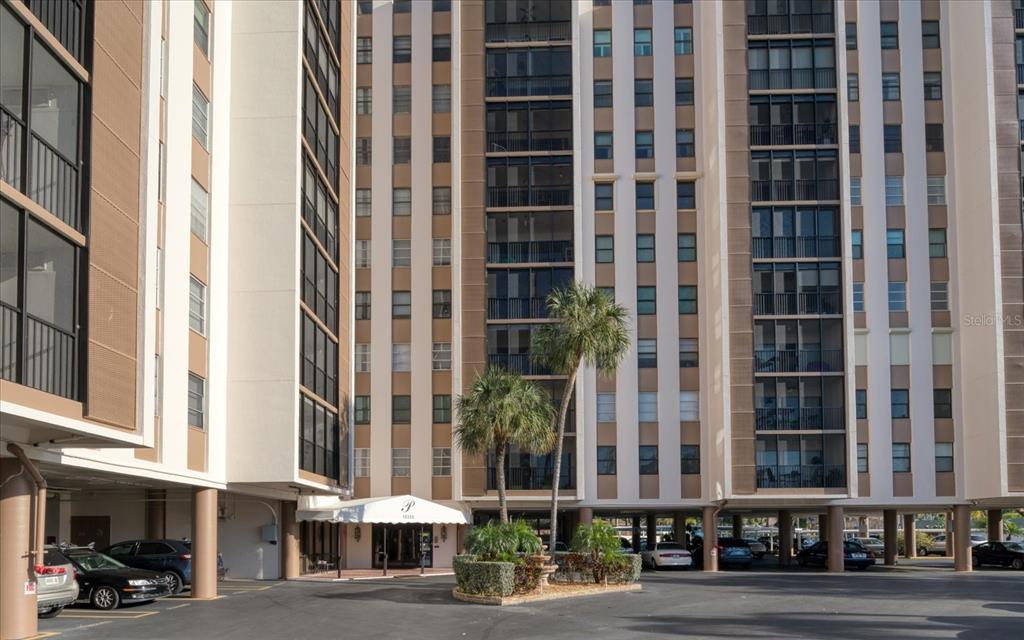 The exterior of a multi-story apartment building features a covered entrance and a parking area — 10355 PARADISE BOULEVARD UNIT 612, TREASURE ISLAND, FL