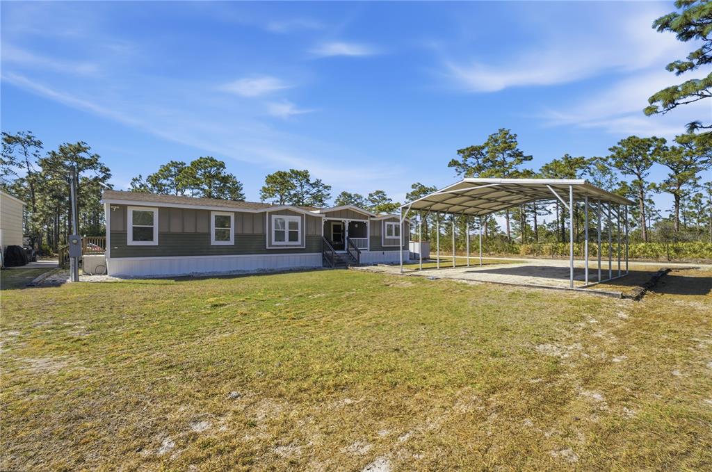A manufactured home with a metal carport sits on a grassy lot with pine trees — 6844 CHAD JASON ROAD, ST CLOUD, FL