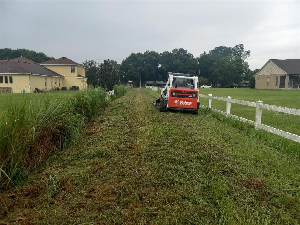 A Bobcat skid-steer mower clears a grassy path between two white fences near residential houses — 3408 N FORBES ROAD, PLANT CITY, FL