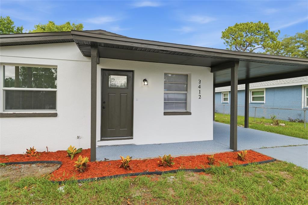 A white house exterior features a dark front door, a carport, and red mulch landscaping — 3412 E HENRY AVENUE, TAMPA, FL