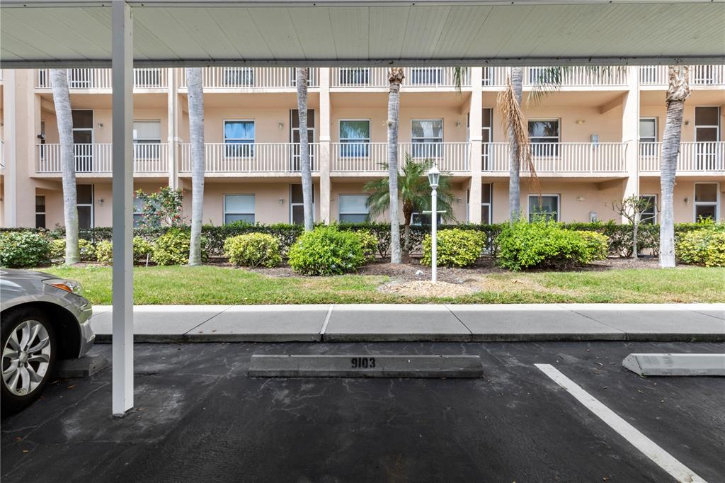 A covered parking space with a numbered concrete bumper sits before a multi-story apartment building — 8750 OLDE HICKORY AVENUE UNIT 9103, SARASOTA, FL