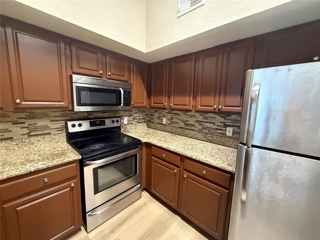 A kitchen features brown cabinets, granite countertops, a tiled backsplash, and stainless steel appliances — 13028 PLANTATION PARK CIRCLE UNIT 1225, ORLANDO, FL