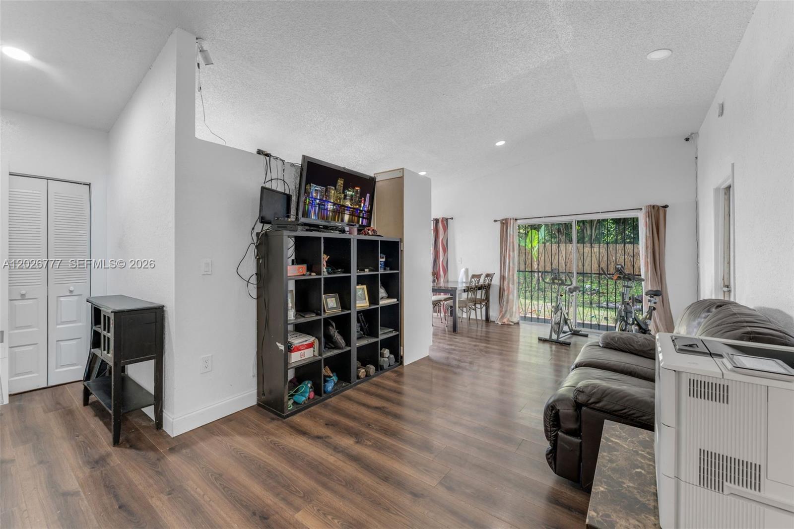 A living room features wood-look flooring, a black shelving unit, and a sliding glass door — 12026 SW 210th St, Miami FL 33177, MIAMI, FL