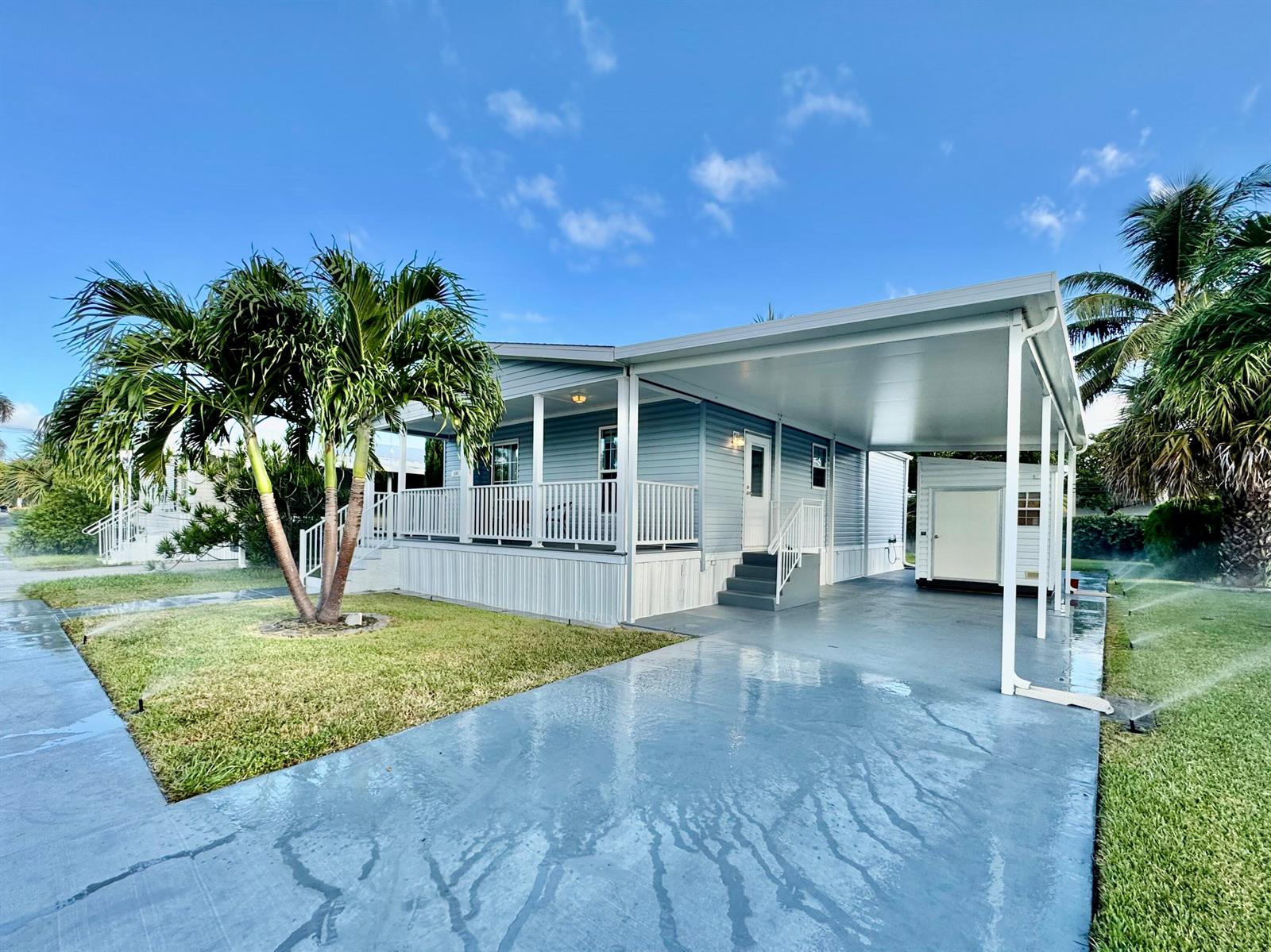 A light blue manufactured home features a covered carport, a front porch, and palm trees — 133 NW 53 Place, Deerfield Beach FL 33064, DEERFIELD BEACH, FL