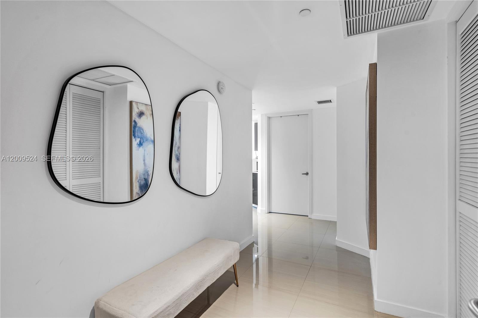 A white hallway features two irregular mirrors, a beige bench, and glossy tile flooring — 31 SE 6th St # 702, Miami FL 33131, MIAMI, FL