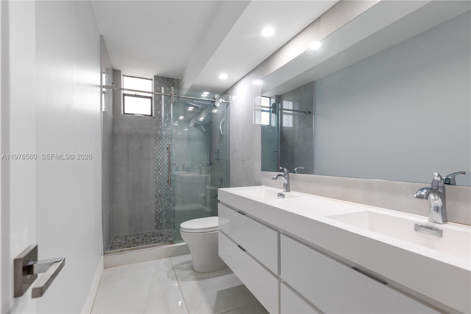 A modern bathroom features a double vanity, a glass-enclosed shower, and light gray tile flooring — 2500 Parkview Dr # 1121, Hallandale Beach FL 33009, HALLANDALE BEACH, FL