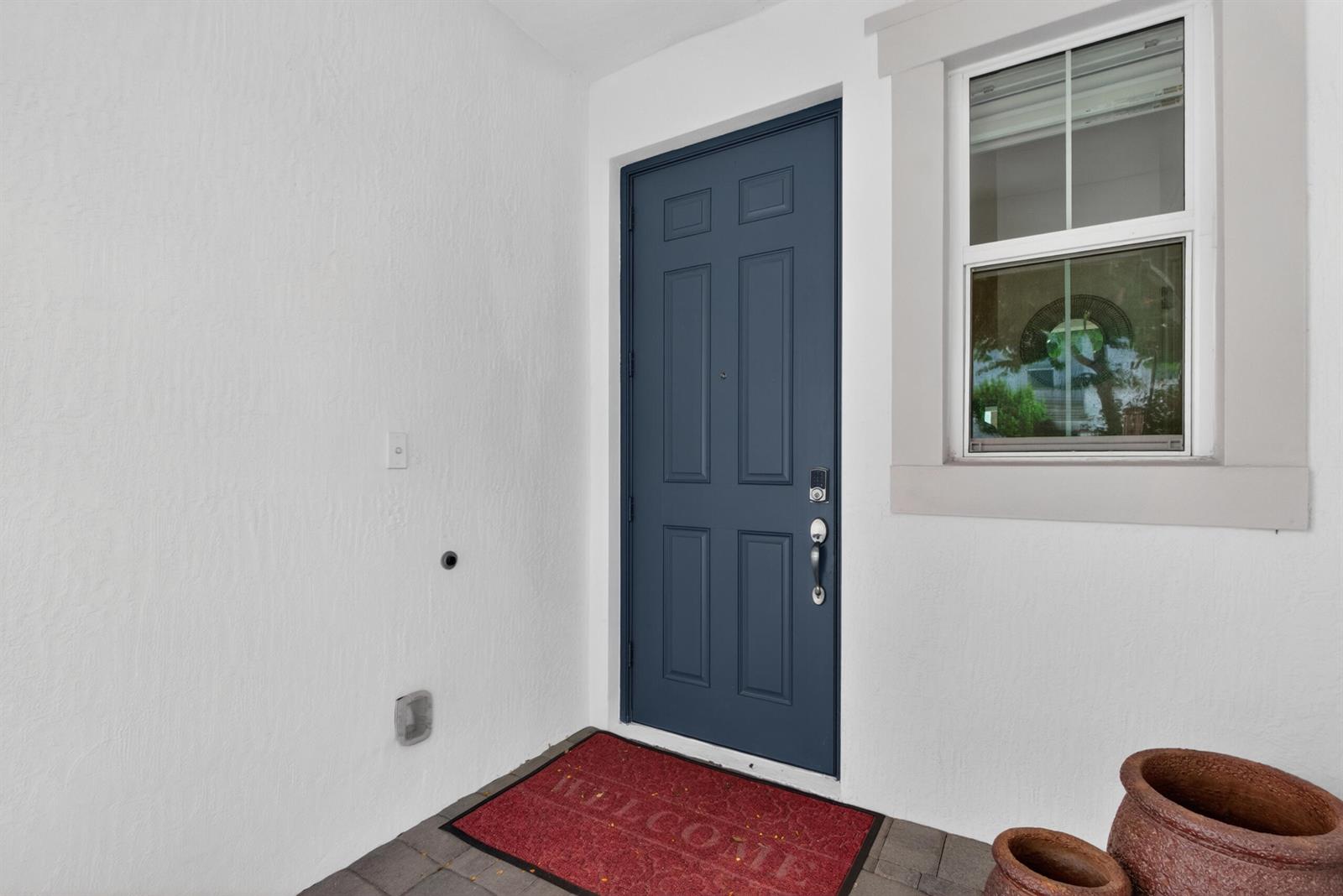 A blue front door with a welcome mat is set in a white exterior wall — 1823 Sandpiper Pointe Place, Deerfield Beach FL 33442, DEERFIELD BEACH, FL
