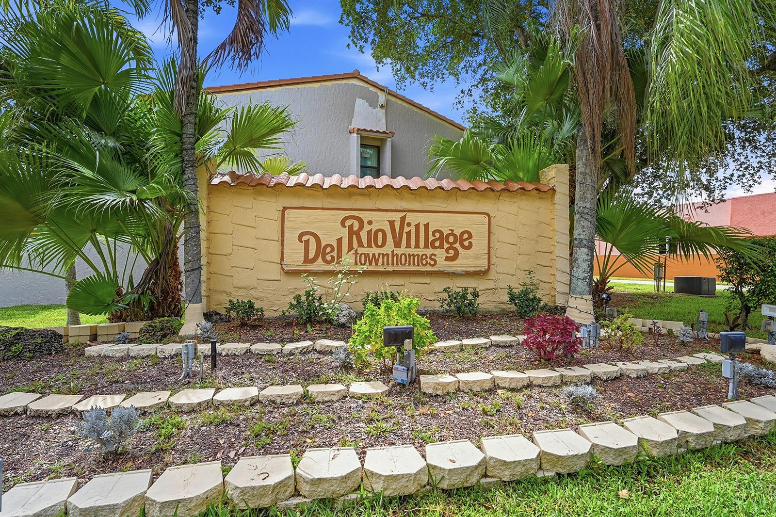 A tan stucco sign for Del Rio Village townhomes sits in front of palm trees — 4012 Del Rio Way, Sunrise FL 33351, SUNRISE, FL