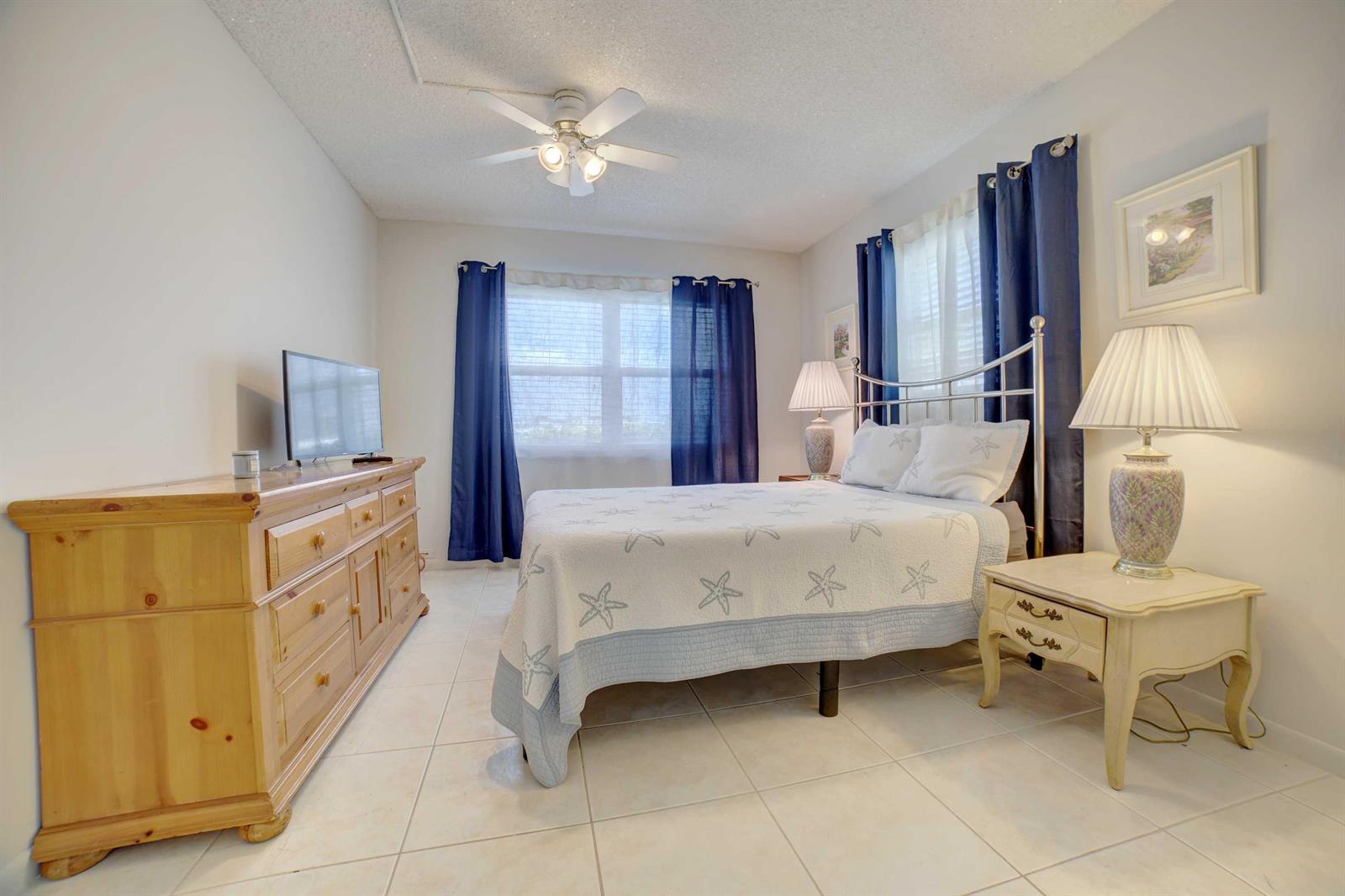 A bedroom with a metal bed frame, wooden dresser, tiled floors, and blue curtains — 1801 Ocean Drive # 110, Boynton Beach FL 33426, BOYNTON BEACH, FL