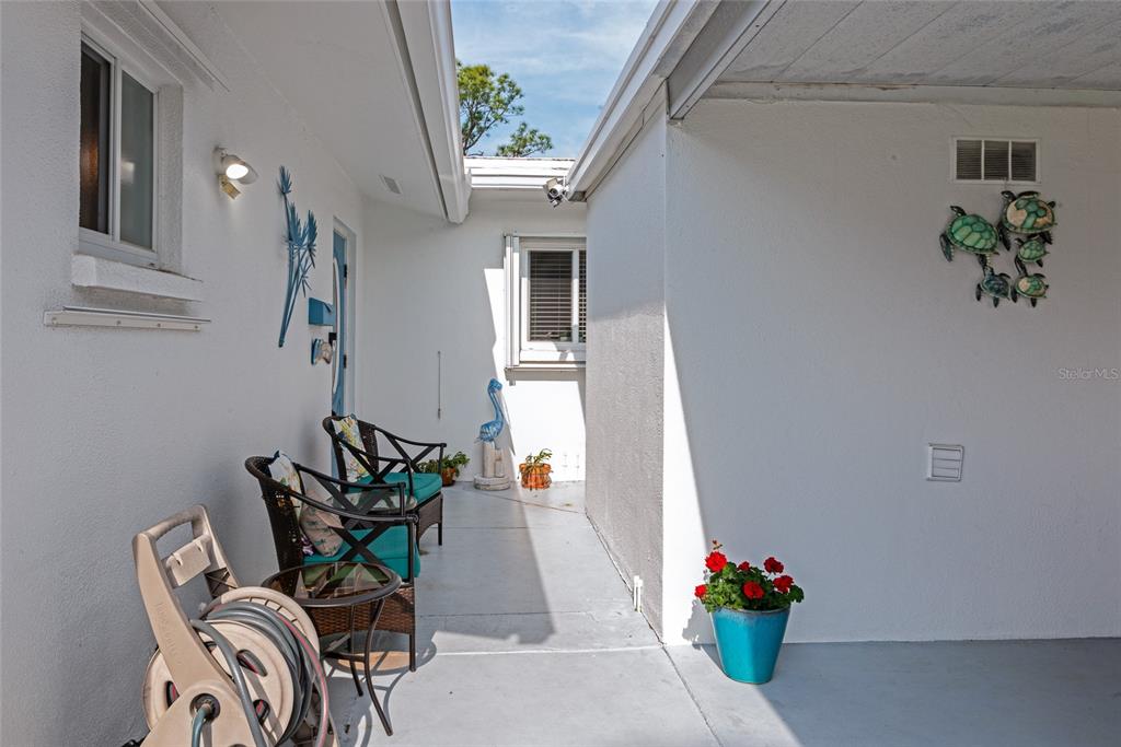 A covered outdoor patio features white walls, two wicker chairs, a hose reel, and decor — 1807 UNIVERSITY PLACE UNIT 151, SARASOTA, FL