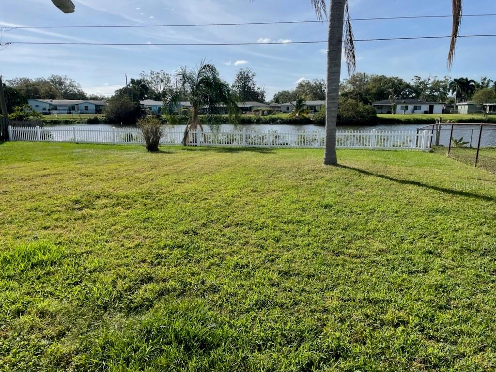 A grassy backyard features a white picket fence overlooking a canal with houses in the background — 5982 52ND AVENUE N, KENNETH CITY, FL