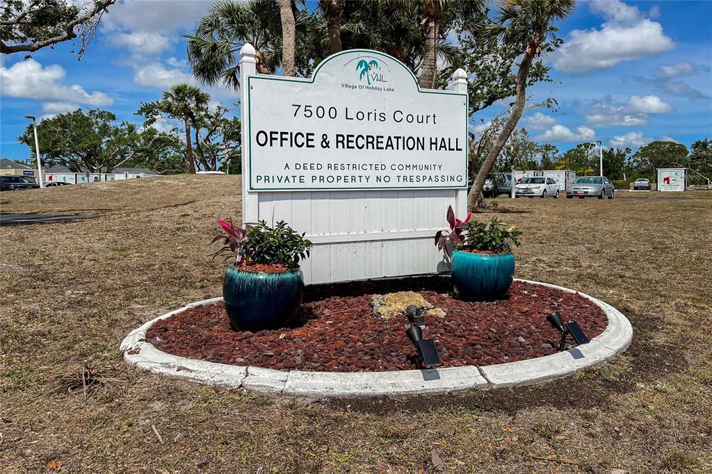 A white sign for a community office and recreation hall stands in a landscaped bed — 7540 DANVERS CIRCLE, PORT CHARLOTTE, FL