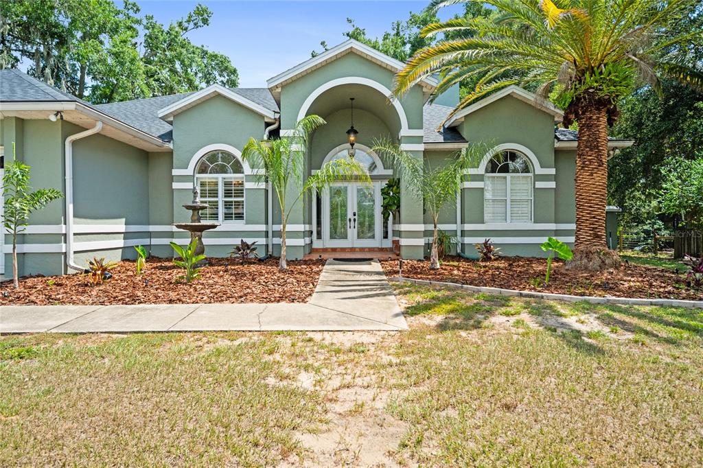 A green single-story house features a central arched entryway, double doors, and a palm tree — 1254 MAGNOLIA FARMS CT, EUSTIS, FL