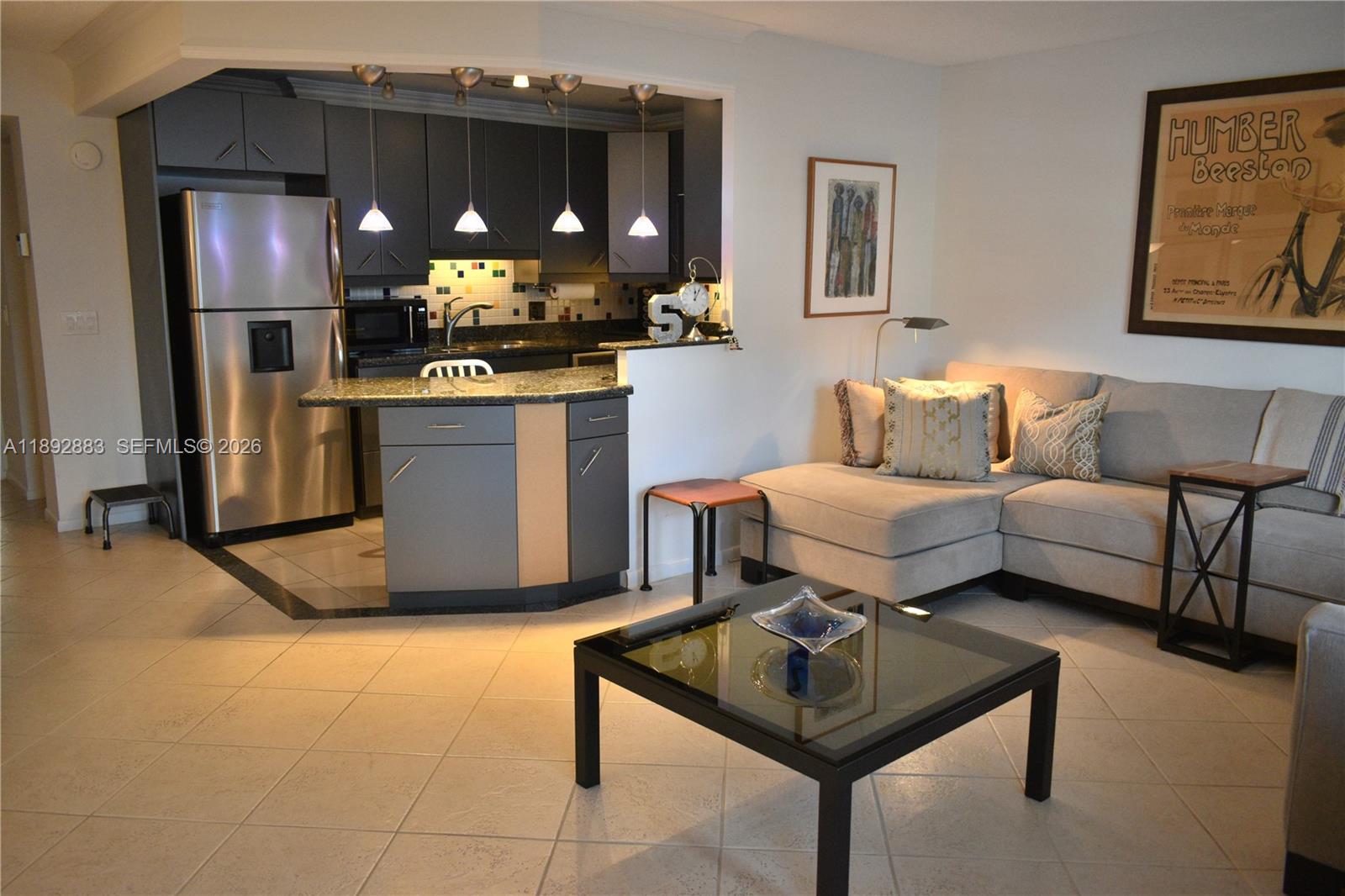 An open-concept living room features a kitchen with dark cabinetry, stainless appliances, and tile flooring — 2011 Cornwall # A, Boca Raton FL 33434, BOCA RATON, FL