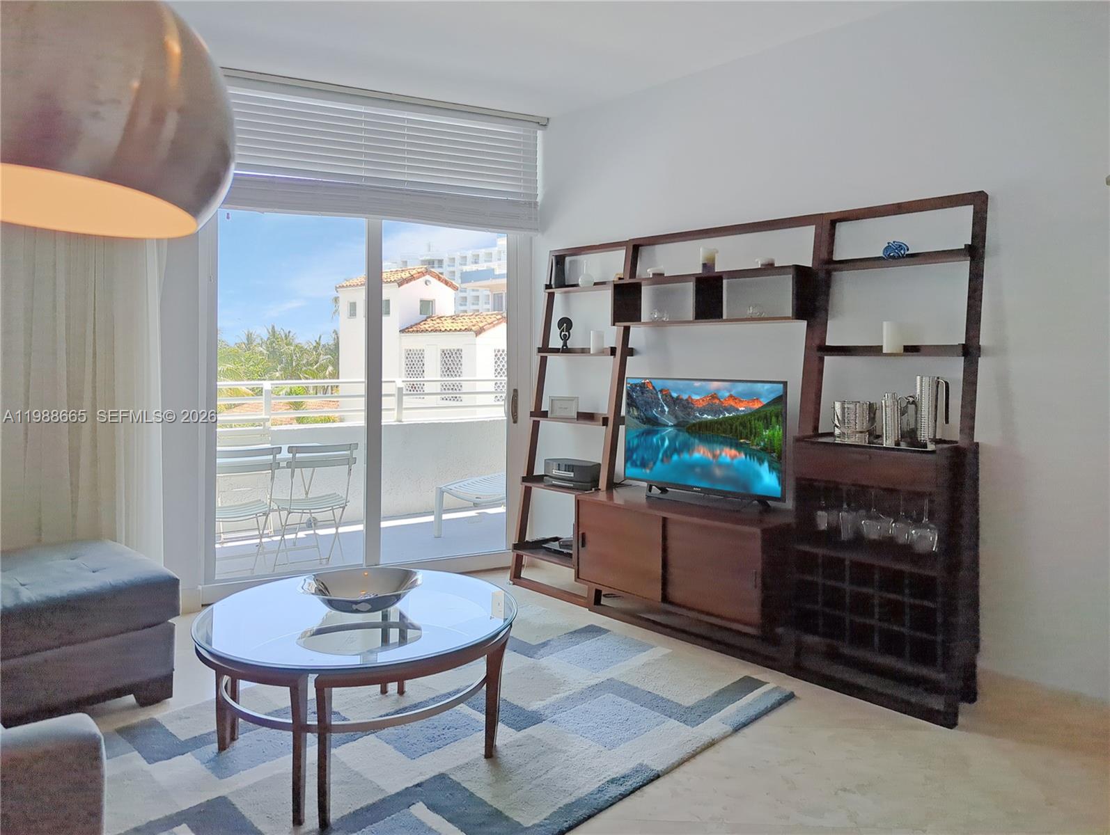 A living room features a glass coffee table, a wooden shelving unit, and a balcony view — 225 Collins Ave # 4J, Miami Beach FL 33139, MIAMI BEACH, FL