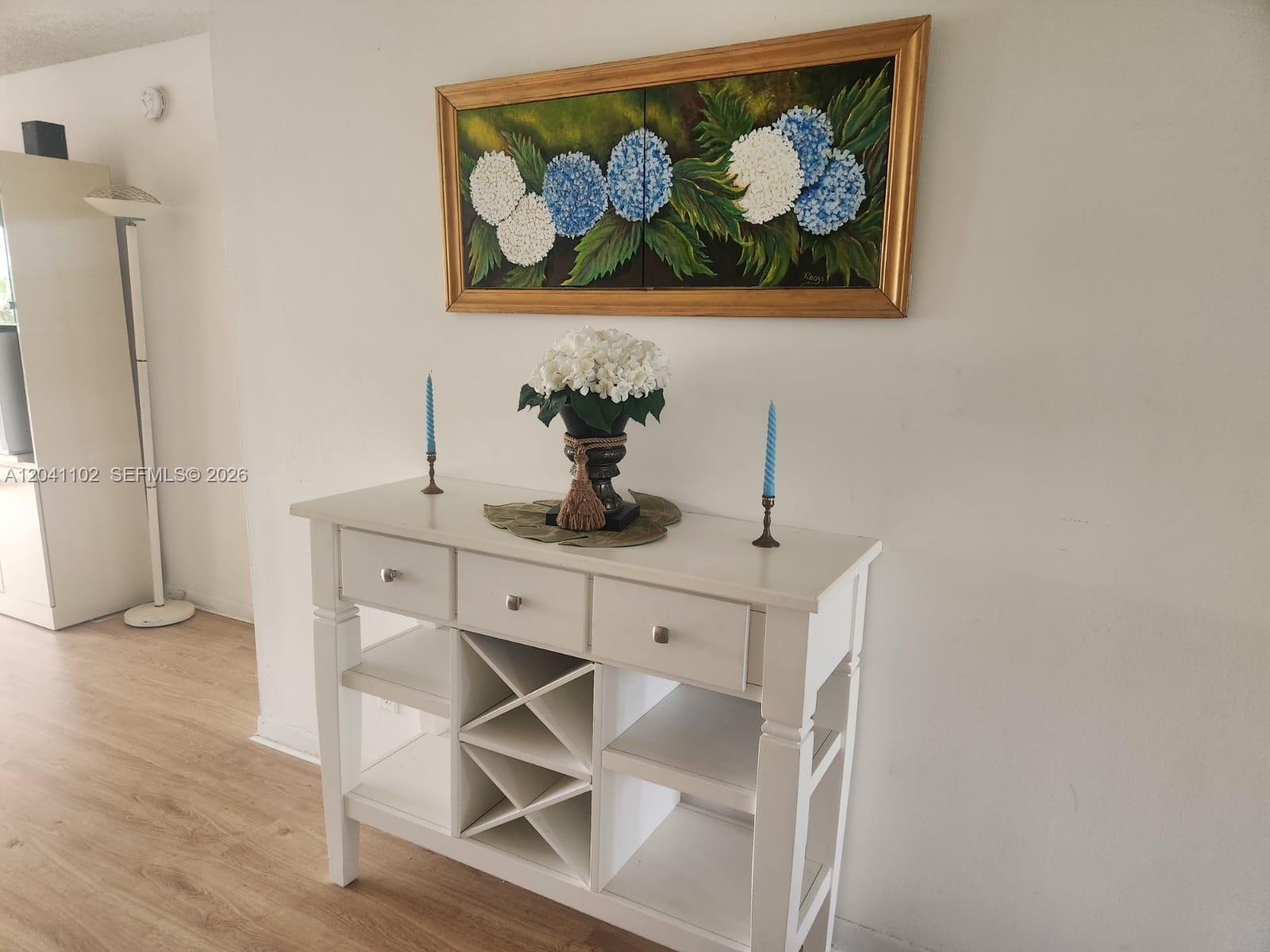 A white console table with drawers and wine storage sits beneath a floral painting — 3571 Inverrary Dr # 307, Lauderhill FL 33319, LAUDERHILL, FL