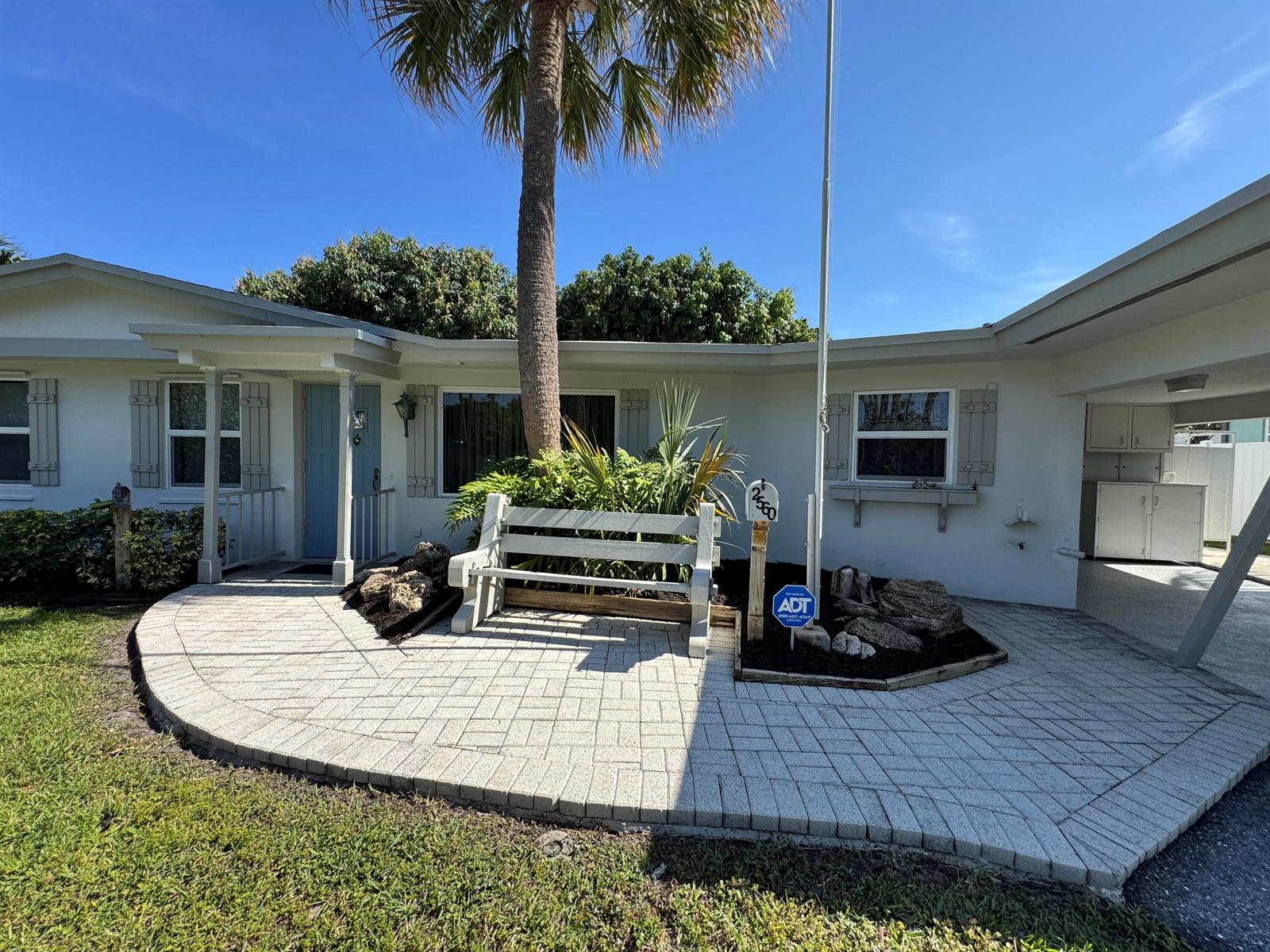 A white house exterior features a paved patio, a wooden bench, and a palm tree — 2560 NE 20th Street, Pompano Beach FL 33062, POMPANO BEACH, FL