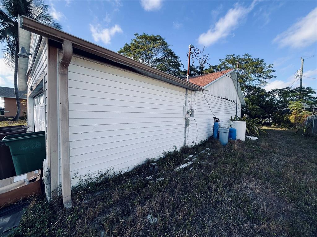 The exterior side of a white-sided house features a gutter, utility equipment, and overgrown grass — 2247 SW 4TH STREET, CAPE CORAL, FL