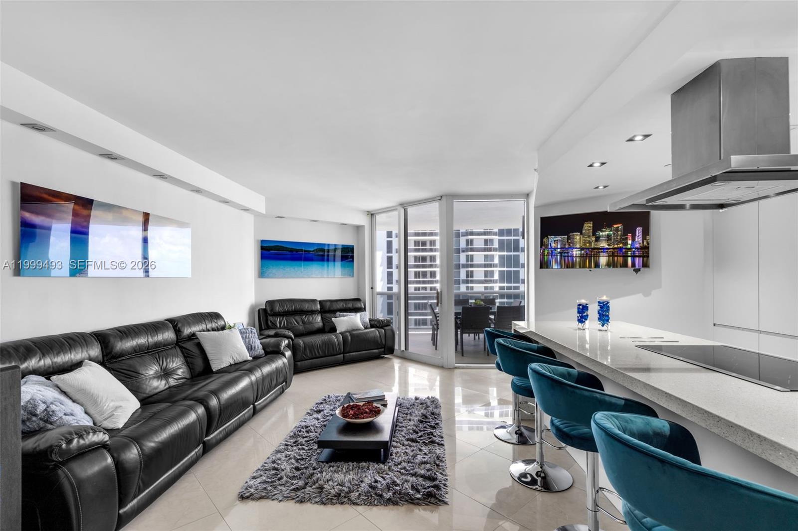 A modern living room and kitchen area features black leather sofas, blue barstools, and balcony access — 4775 Collins Ave # 3608, Miami Beach FL 33140, MIAMI BEACH, FL