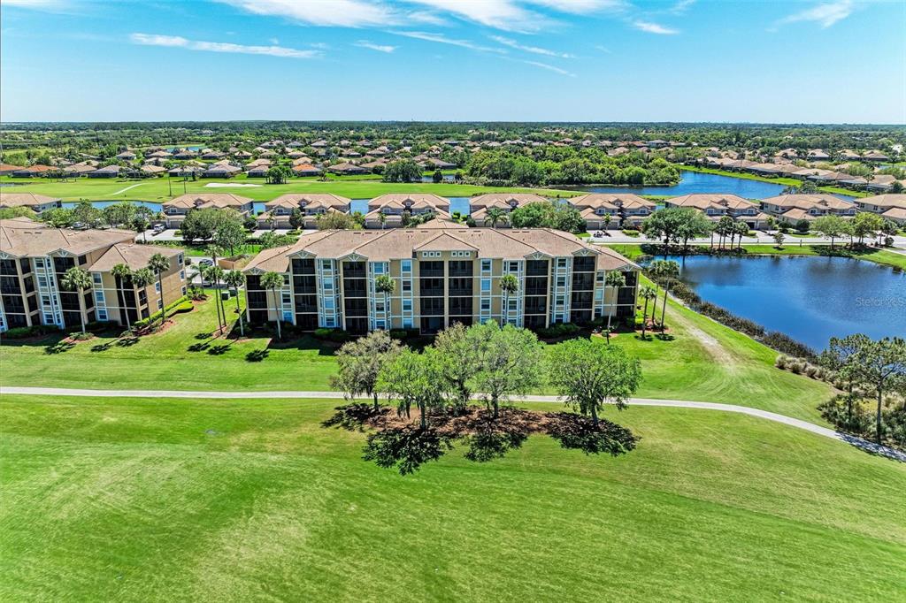 An aerial view shows a multi-story apartment building situated beside a lake and golf course — 8105 GRAND ESTUARY TRAIL UNIT 403, BRADENTON, FL