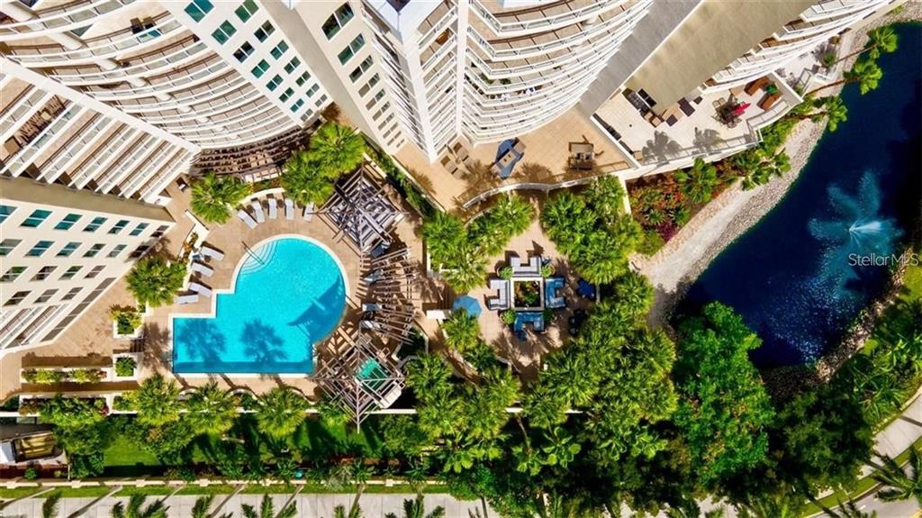 An aerial view shows a resort-style swimming pool, patio area, and pond beside tall buildings — 1300 BENJAMIN FRANKLIN DRIVE UNIT 301, SARASOTA, FL