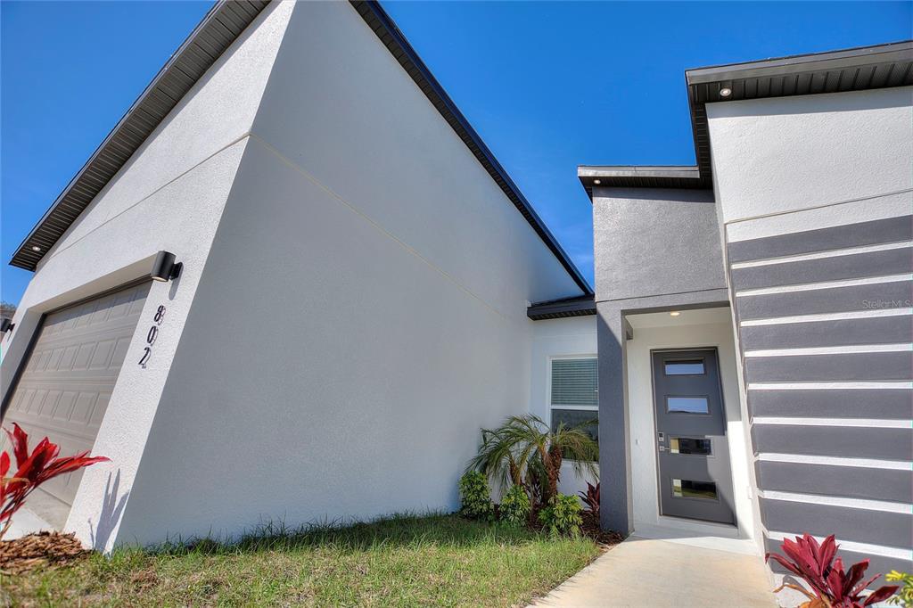 A modern house exterior features a gray garage door, a front door, and landscaping — 802 JAMES DRIVE, POINCIANA, FL
