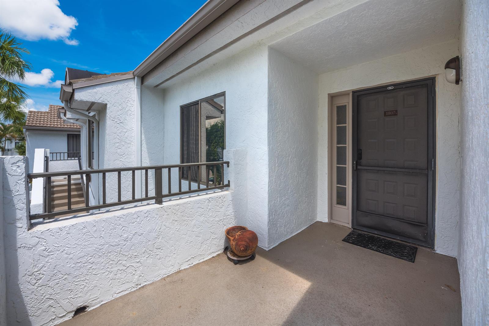 A white stucco exterior entryway features a brown door, a metal railing, and a seashell planter — 7689 Glendevon Lane # 1807, Delray Beach FL 33446, DELRAY BEACH, FL