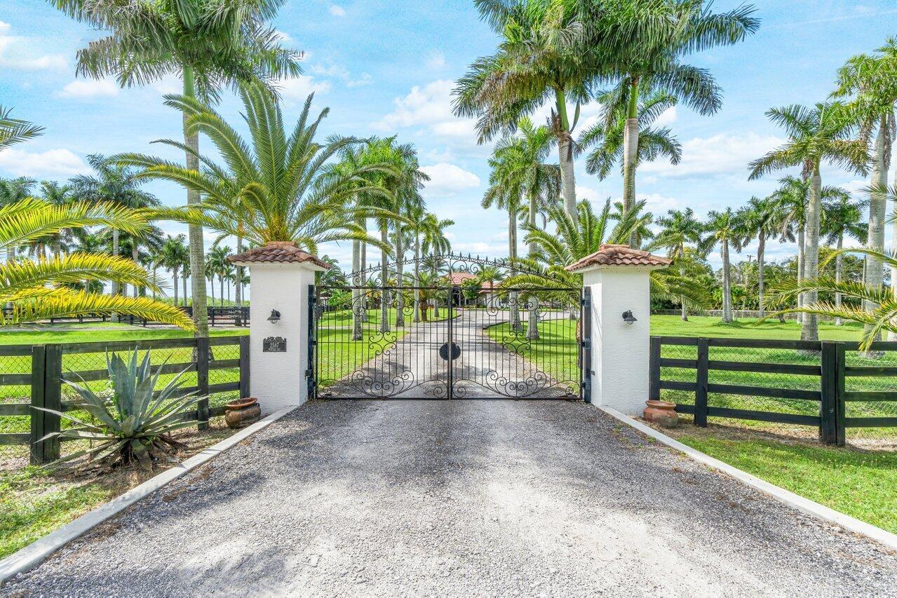 A gravel driveway leads to a gated entrance flanked by white pillars and palm trees — 2587 Skipiks Way, Wellington FL 33470, WELLINGTON, FL