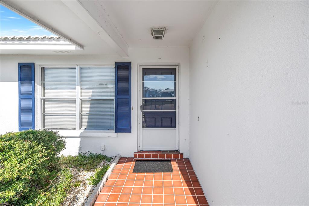 A white exterior wall with a tiled entryway, a storm door, and blue-shuttered windows — 802 BUTTONWOOD DRIVE, BRADENTON, FL
