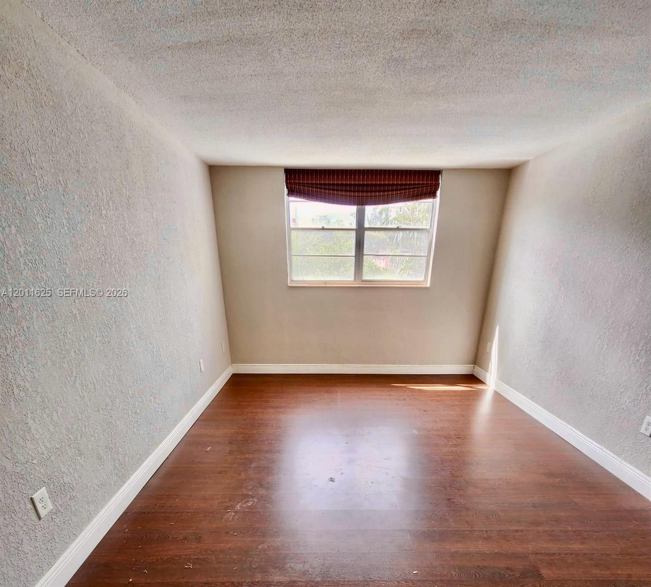 An empty room features textured walls, dark wood-look flooring, and a window with a valance — 12590 NE 16th Ave # 209, North Miami FL 33161, NORTH MIAMI, FL